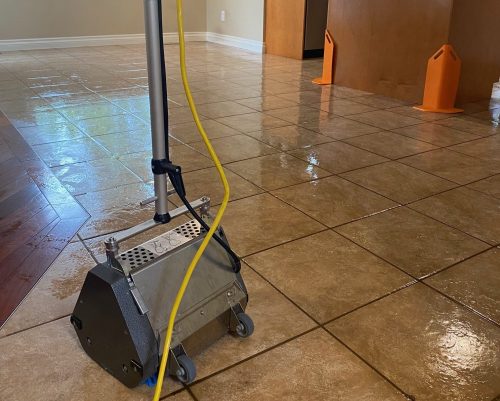 A commercial floor scrubbing machine sitting on wet, freshly cleaned ceramic tile floor in a residential kitchen area, with orange safety signs placed nearby, highlighting professional tile, grout, and hard-surface floor cleaning services.