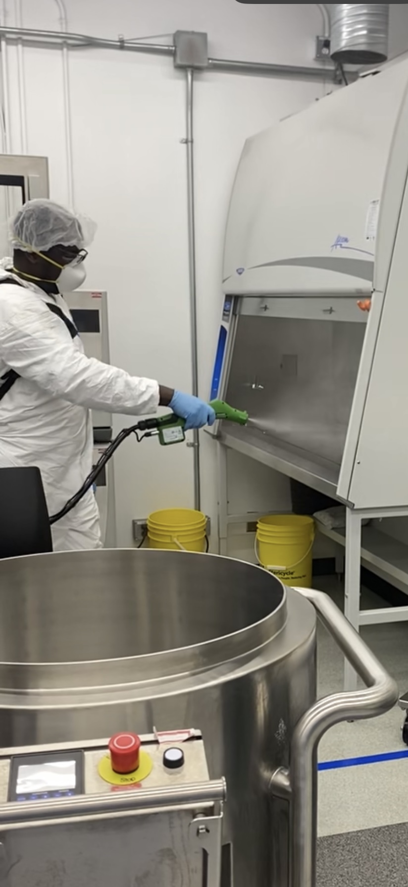 A professional sanitation worker in full protective personal equipment (PPE), including a suit, mask, and gloves, using a specialized spray wand to sanitize a biosafety cabinet inside a laboratory, showcasing commercial electrostatic spraying and medical decontamination services.