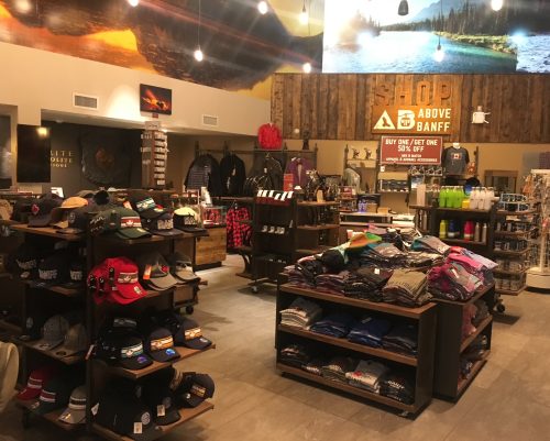 A well-organized retail souvenir shop featuring neatly arranged displays of baseball caps, folded apparel, water bottles, and gift merchandise on dark wood shelving, with clean tile floors under warm ceiling lights, highlighting commercial retail cleaning and store janitorial services.