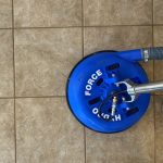 A top-down view of a blue Hydro-Force tile and grout cleaning tool operating on a textured tan ceramic tile floor, highlighting professional hard-surface floor cleaning and grout restoration services.