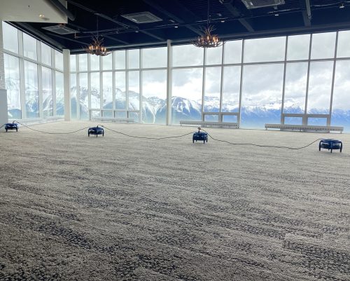 A large, open event space with wall-to-wall carpet and floor-to-ceiling windows offering a panoramic view of snow-capped mountains. Four commercial carpet drying fans are set up across the room, connected by power cords, highlighting professional commercial carpet cleaning services.