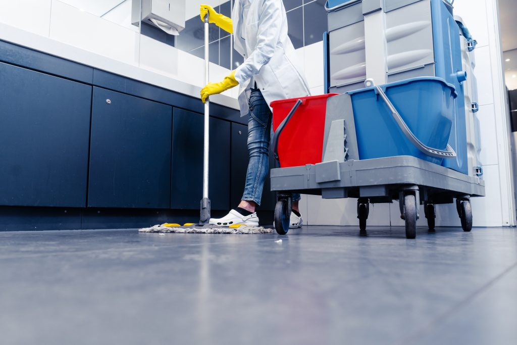 A low-angle view of a cleaning professional wearing yellow gloves mopping a dark tiled floor next to a fully equipped janitorial cleaning cart, highlighting commercial floor maintenance and janitorial services.